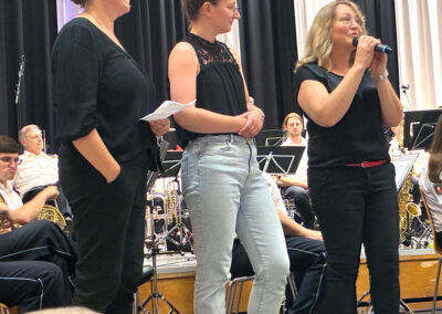 Die Organisatorinnen stehen vor dem Orchester und sprechen in ein Mikofon
